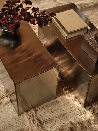 Two rectangular metallic side tables with a brushed bronze finish, styled on a textured beige shag rug with a book and a plant in the background.