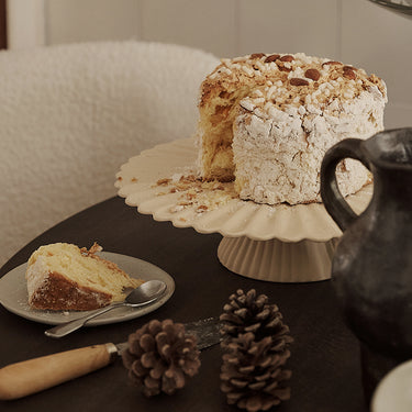 Interior image of a cake stand with a large cake with a slice removed on a dark wooden table.