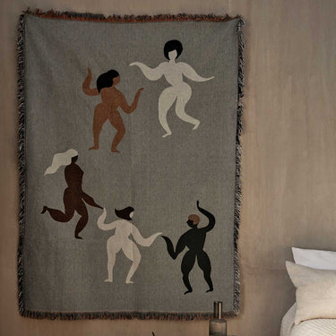 A tapestry blanket with a motif of five dancing figures in shades of beige, brown, and black, with decorative fringing along the edges, displayed on a sofa.