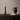 Interior image of a black brass candle holder with a candle in it. On a dark stained wooden cabinet with a water jug, a book and glass of water.