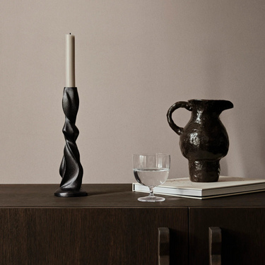 Interior image of a black brass candle holder with a candle in it. On a dark stained wooden cabinet with a water jug, a book and glass of water.