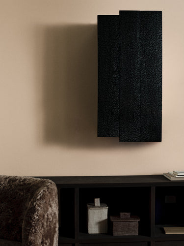 A modern black textured wall art piece with three overlapping vertical panels, mounted on a beige wall above a dark console table.