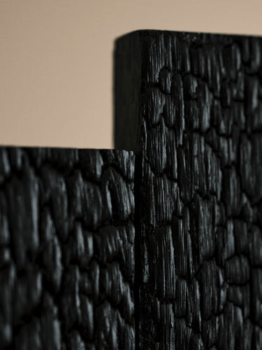 Close-up of a black textured wall panel with a cracked, charred wood-like finish, shown against a warm beige background.