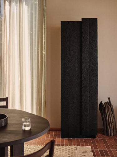 A tall black textured room divider screen standing in a warm-toned interior next to a window with sheer curtains, with a round wooden table and chairs in the foreground.