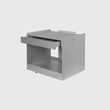 A minimalist, modern side table or nightstand made of brushed metal, featuring a single pull-out drawer and an open lower shelf, shown on a light gray background.