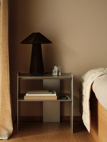 A modern two-tier metal nightstand in a warm-toned bedroom setting, styled with a dark woven table lamp, a small glass, and books, positioned next to a bed with a cream quilt.