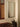 A tall, narrow rectangular wall mirror with a slim silver frame, mounted on a beige wall in a styled interior setting with a wooden bench and decorative trunk.
