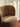 A close-up of a modern barrel-style armchair upholstered in a textured taupe-brown fabric, set in a room with warm sunlight streaming through sheer white curtains and a terracotta tile floor visible in the corner.