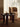 A dark round wooden coffee table with three thick legs in a warm, sunlit room next to a beige upholstered armchair. On the table are a glass of water, a small decorative bowl, and a dark ceramic object, all resting on a brown woven rug.
