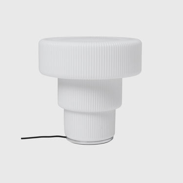 A modern white table lamp with a ribbed, tiered cylindrical design, shown against a light gray background with its black power cord visible on the left.