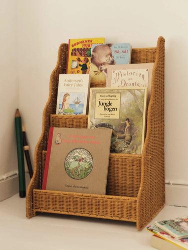 A tall, woven wicker bookshelf with three open compartments holding several children's books, photographed in a real indoor setting against a white wall with pencils on the floor nearby.