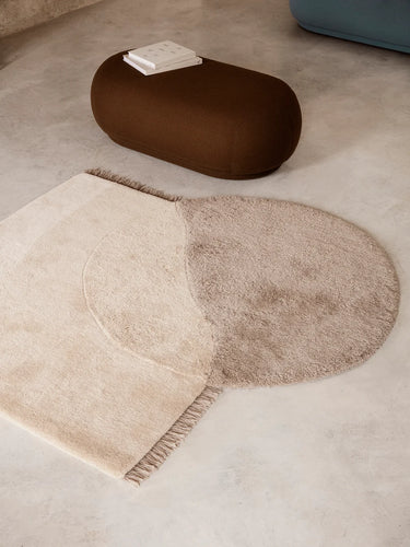A modern area rug in a two-tone design of cream and taupe, styled on a light concrete floor next to a brown pouf.