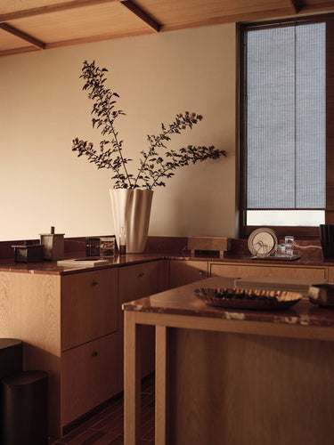 A warm, minimalist interior scene with light wood cabinetry, a marble-topped island, and a tall white vase holding leafy branches against a beige wall. A window with a dark mesh screen is visible on the right.