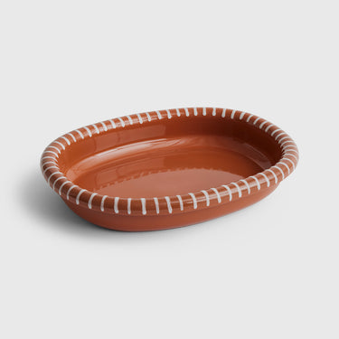 An oval ceramic serving platter in a warm terracotta orange-brown glaze, decorated with a white stitched-look pattern around the rim, shown on a light gray background.