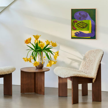 A contemporary walnut lounge chair with a beige upholstered backrest and seat, positioned in a room with a painting, a vase of yellow flowers, and an area rug.