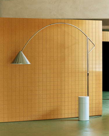 An elegant modern floor lamp with a sweeping curved chrome arch and a white cylindrical base, positioned in front of a warm orange tiled wall.