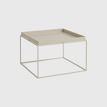 A minimalist square side table featuring a light beige tray on top of a thin, open-frame metal base in a matching beige tone, set against a solid light gray background.