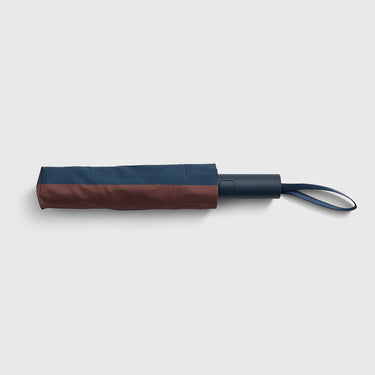 A folded compact umbrella with a two-tone design, featuring a dark navy blue top section and a brown bottom section, with a matching dark navy handle and loop on a light gray background.