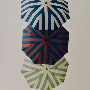 Three striped umbrellas arranged vertically against a plain light background. The top umbrella has blue and white stripes, the middle has navy and muted red stripes, and the bottom has pale green and cream stripes.