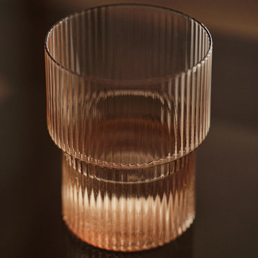 A clear ribbed glass tumbler with a wide upper rim and a shorter lower section, shown on a dark reflective surface under warm lighting.