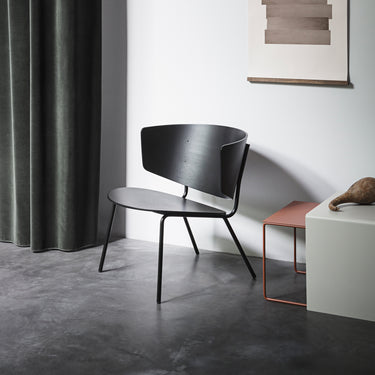 A black Herman Lounge Chair with a steel frame and natural oak veneer seat and backrest, placed on a grey floor next to a small round side table with a decorative item on top.