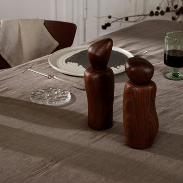 A set of brown wooden cairn grinder with a sculpted design standing upright on a dining table with a stone coloured table cloth.